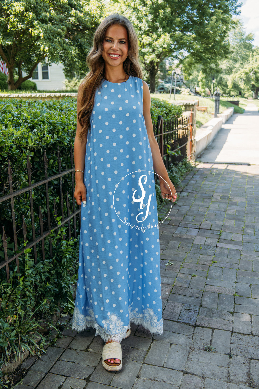 Woman in a blue polka dot dress standing on a sidewalk with greenery in the background