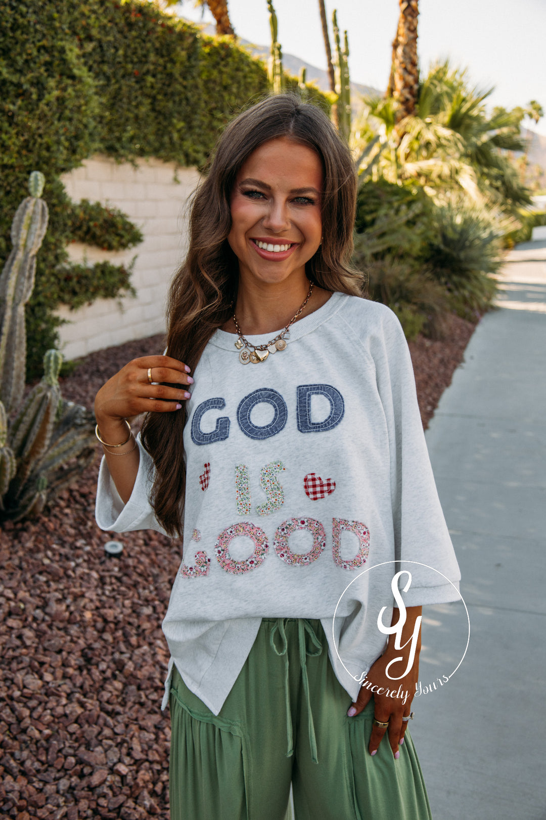 Woman wearing a 'God is Good' shirt outdoors with plants and a path in the background