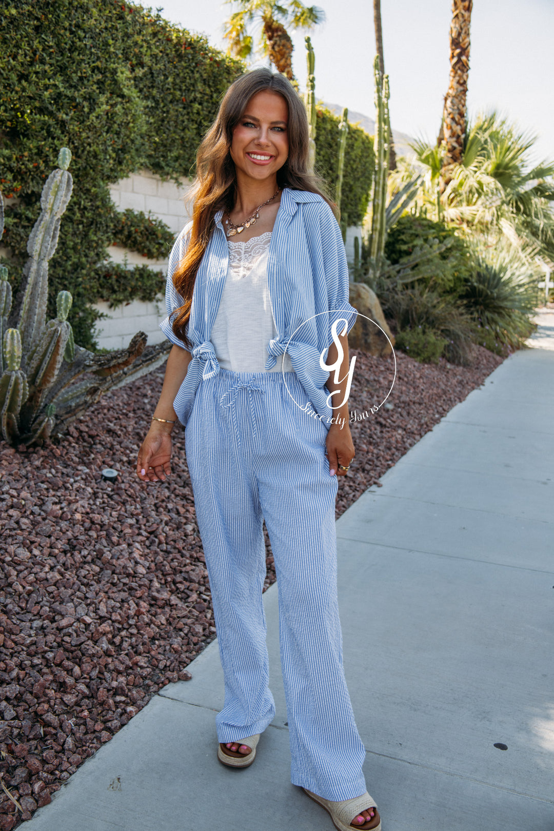 Woman in a light blue outfit standing on a sidewalk with desert plants in the background