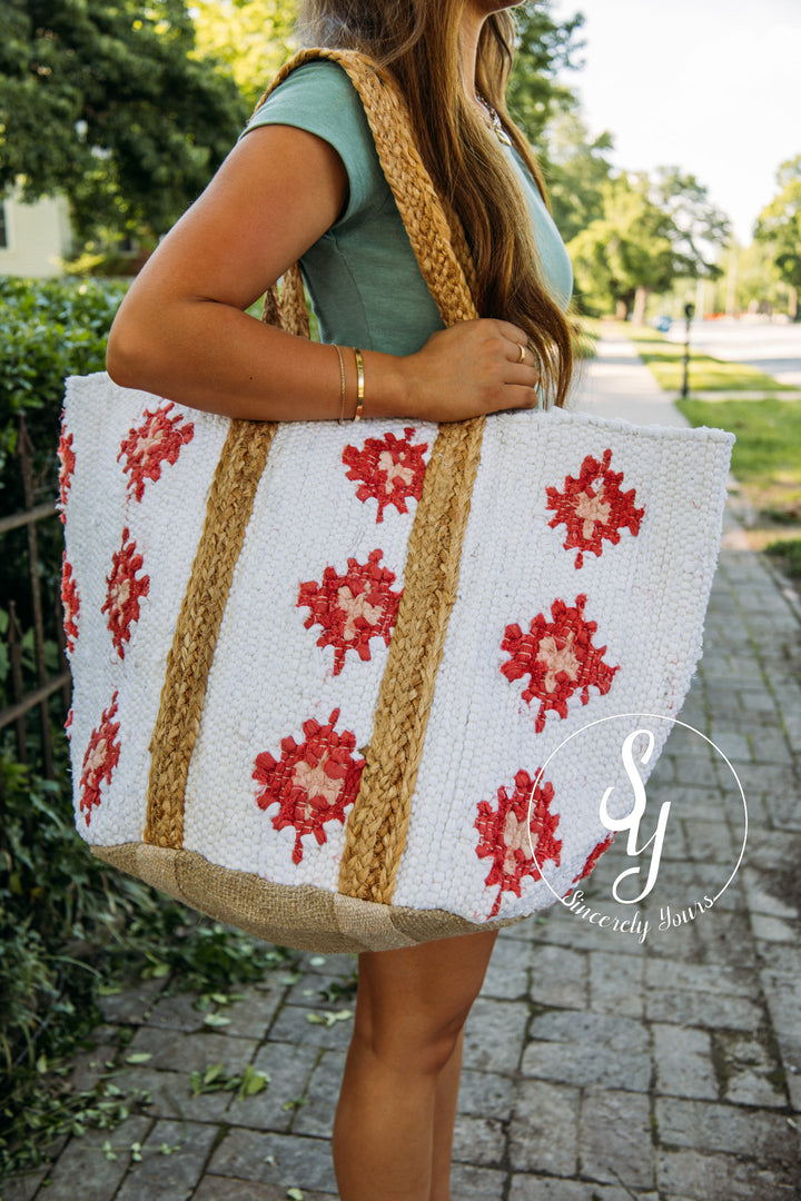 Person holding a white tote bag with red floral patterns and jute material outdoors.