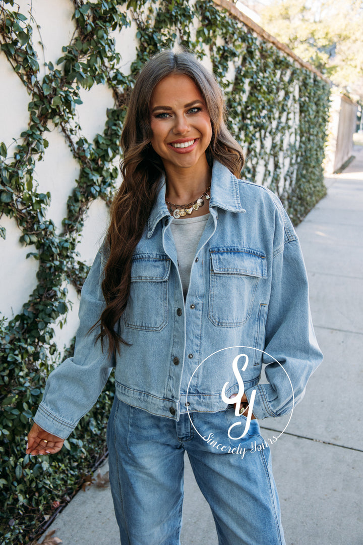 Woman wearing a denim jacket and jeans standing against a wall with greenery