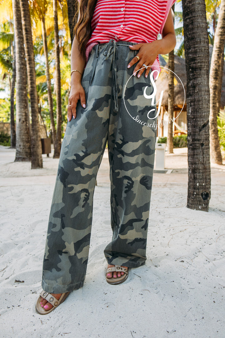 Person wearing camouflage pants and a pink top standing on a sandy beach with palm trees in the background.