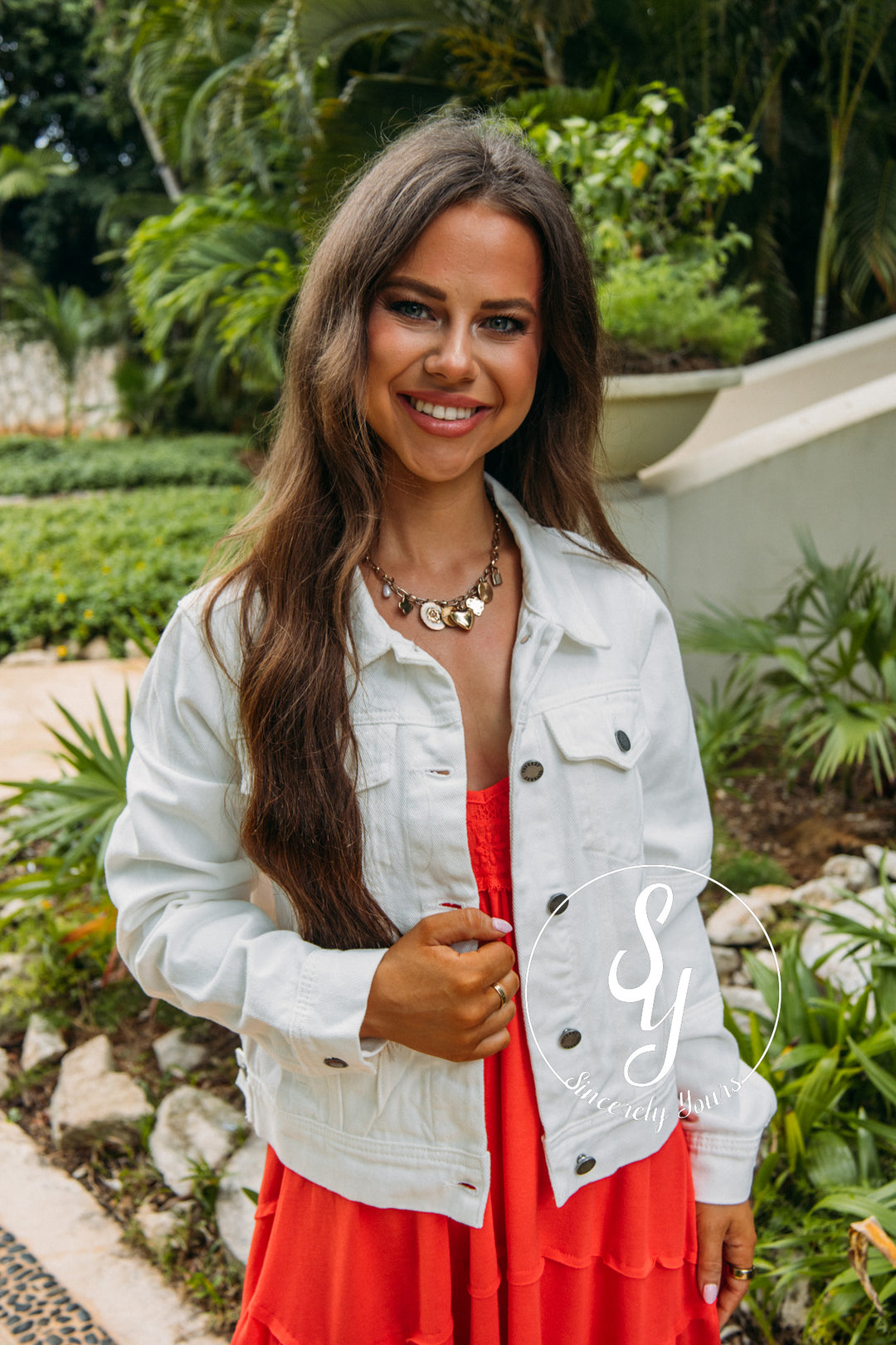 Woman in a white jacket and red dress standing outdoors with greenery in the background