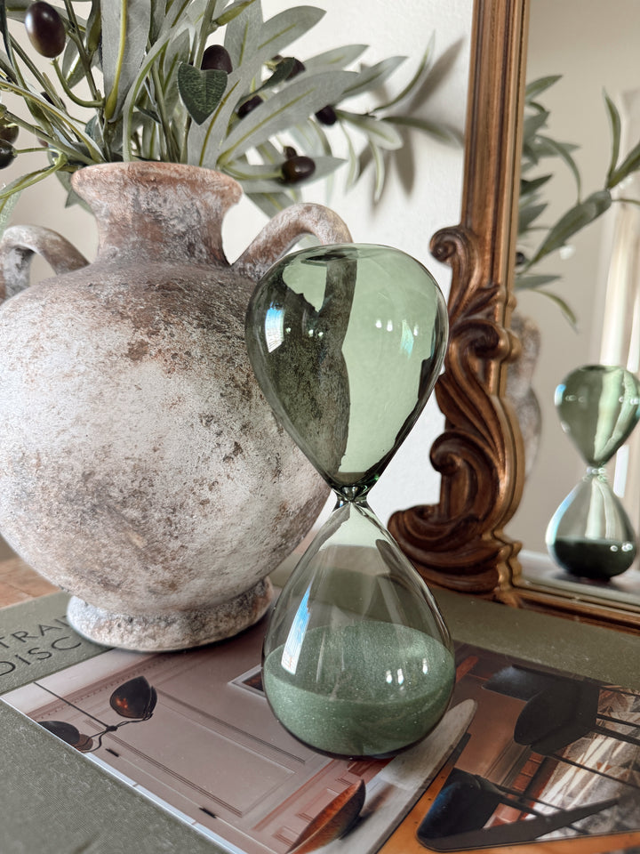 Green hourglass on a surface with a textured vase and decorative mirror in the background